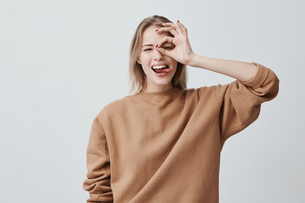 Cheerful excited female with blonde long straight hair showing Ok gestures with both hands, pretending to wear spectacles, and smiling broadly, enjoying her carefree happy life