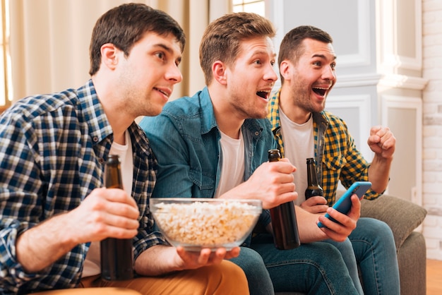 Cheerful excited friends holding beer bottles in hand watching match on television