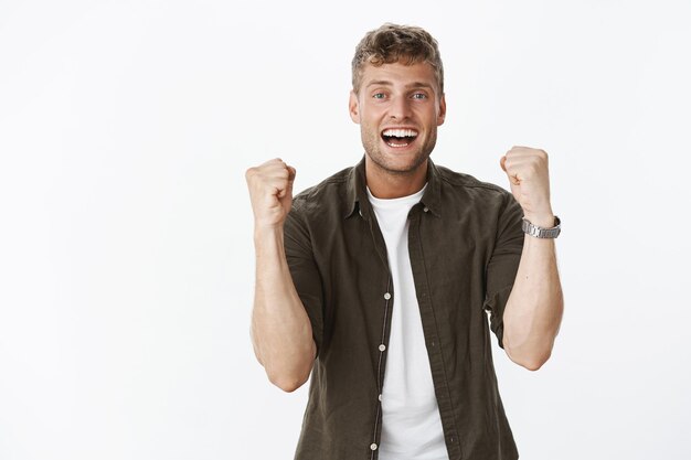 Cheerful excited happy attractive blond guy with bristle and white teeth raising fists joyfully, smiling satisfied triumphing, celebrating victory or success, reacting to awesome news over gray wall.