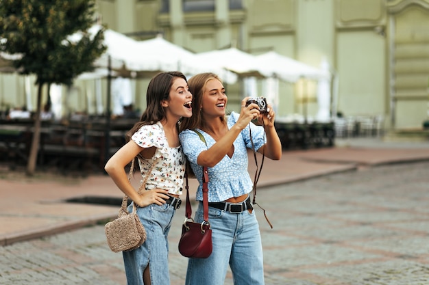 Веселые возбужденные загорелые девушки в стильных блузках с цветочным рисунком и джинсовых штанах искренне улыбаются на улице