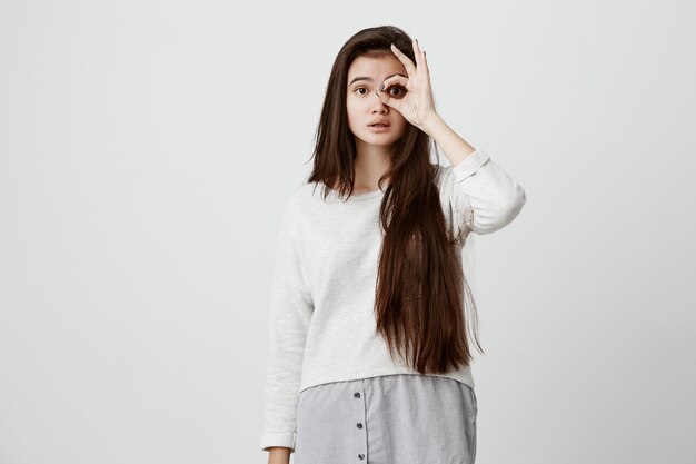 Cheerful excited teenage woman with dark long straight hair showing Ok gesture with hand, enjoying her carefree happy life, dressed casually. Body language and gestures concept.
