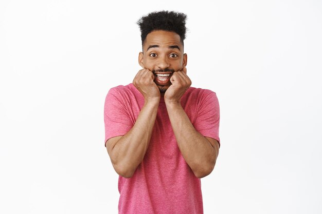 Cheerful and excited young african american guy, squeeze his cheeks and smiling from anticipation and excitement, hear amazing news, waiting hopeful, white background