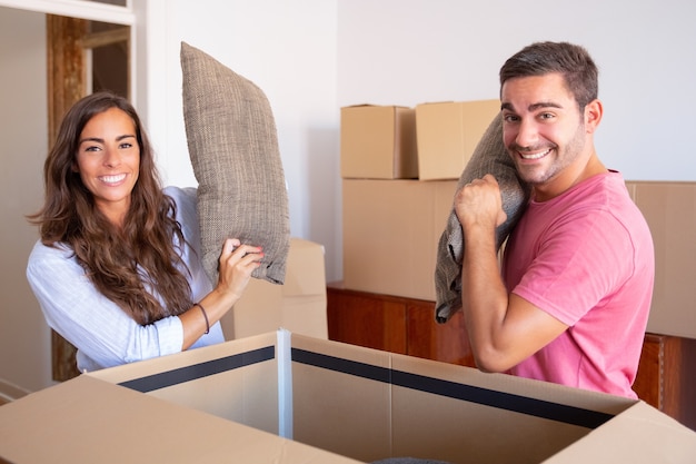 Cheerful excited young man and woman getting out cushions of open carton box, enjoying moving and unpacking things