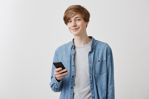 スマートフォンでインターネットを閲覧するダニムシャツで陽気な金髪の白人男性学生。現代の技術とコミュニケーション