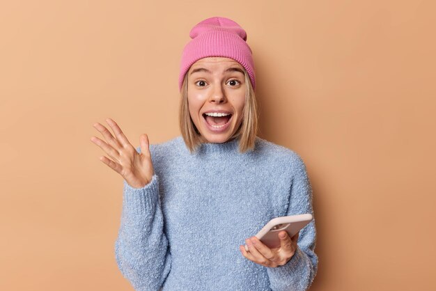 Cheerful fair haired young European woman keeps hand raised exclaims loudly uses smartphone for chatting online wears hat and casual jumper isolated over brown background. Human emotions concept