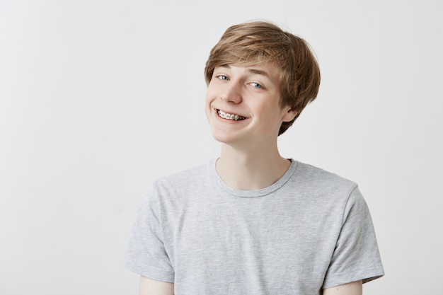 Free photo cheerful fair-haired young man with braces grins , has overjoyed expression, clenches teeth, being glad to pass all exams at university. positive feelings and emotions.