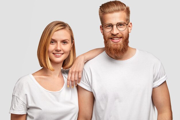 Cheerful family couple rejoice they become parents soon, stad close to each other, dressed in casual clothes, isolated over white wall. Happy bearded male has date with pretty young female