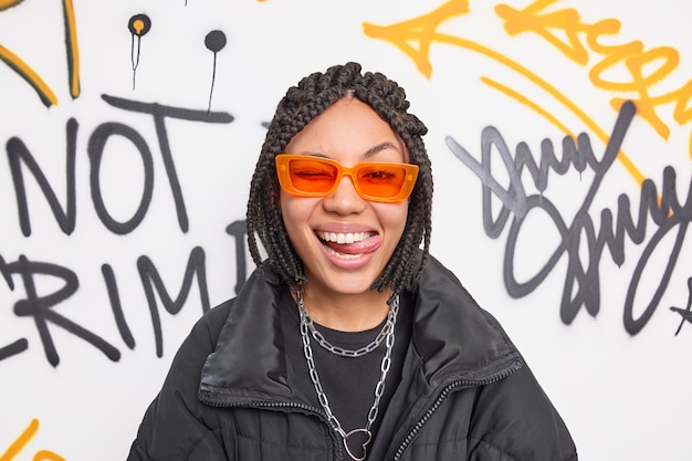 Free photo cheerful fancy teenage girl wears trendy orange suglasses and black jacket sticks out tongue has dreadlocks hairstyle expresses positive emotions poses against graffiti wall