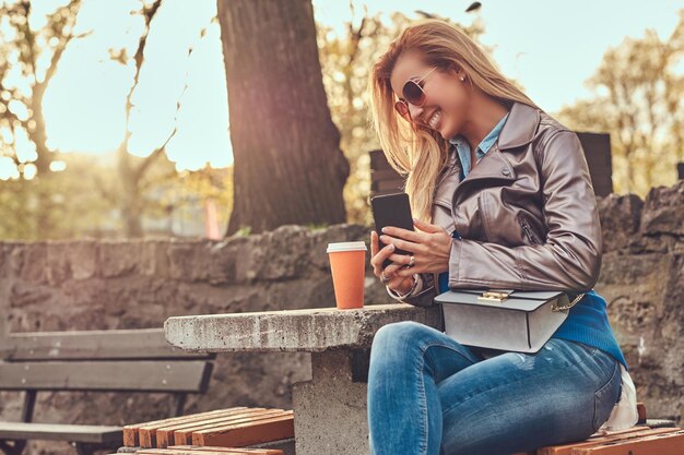 Cheerful fashionable blonde blogger female relaxes outdoor, using a smartphone while sitting on the bench in city park against a bright sunlight.