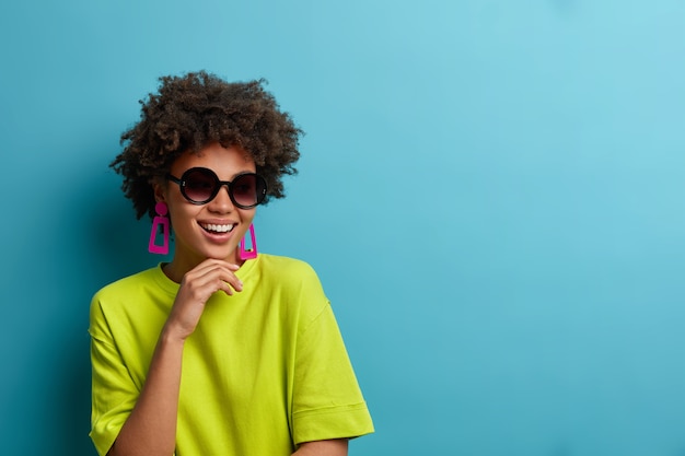 Cheerful fashionable curly ethnic woman holds chin, wears trendy sunglasses and green t shirt, has happy summer mood, poses for magazine cover, isolated on blue wall with copy space aside