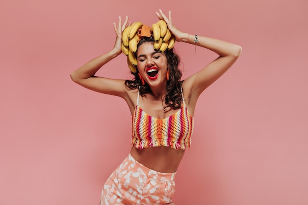 Cheerful fashionable lady with wavy hairstyle and orange accessories in bright summer clothes rejoices and holding bananas..
