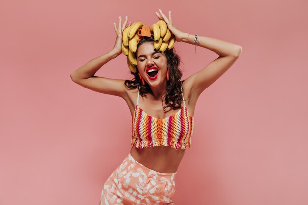 明るい夏の服を着た波状の髪型とオレンジ色のアクセサリーを持つ陽気なファッショナブルな女性は、バナナを持って喜んでいます。