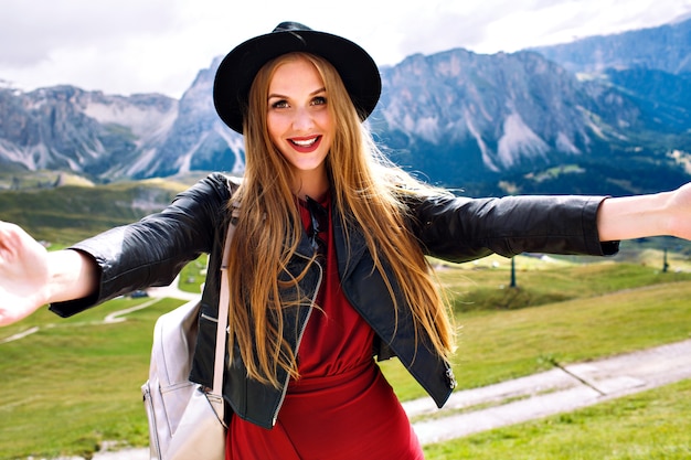 Cheerful fashionable young model woman making selfie at Alp mountains, wearing dress, leather jacket, sunglasses and backpack
