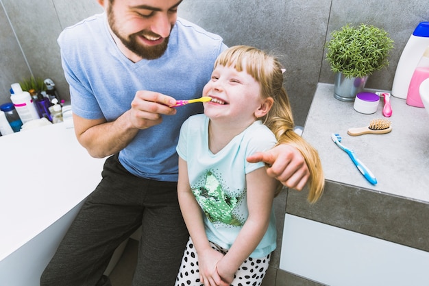 Cheerful father helping daughter to brush teeth