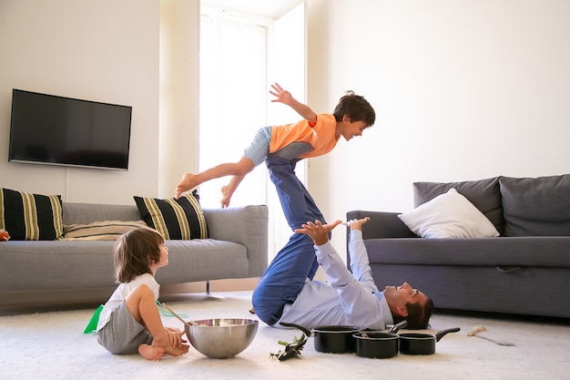 Cheerful father holding son on legs and lying on carpet. Happy Caucasian boy flying in living room with help of dad. Cute boy sitting on floor near bowl and pans. Childhood and weekend concept