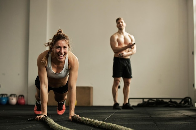 Бесплатное фото Веселая спортсменка отжимается, тренируясь с боевой веревкой и развлекаясь в спортзале. на заднем плане мужчина.