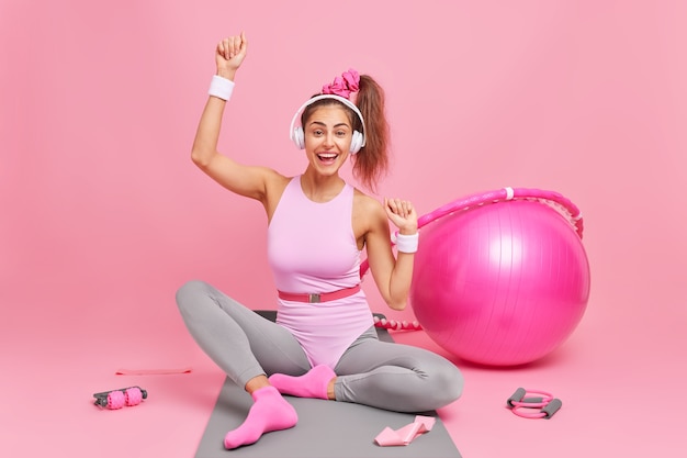 Cheerful female athlete with pony tail in activewear enjoys favorite playlist via wireless headphones poses on fitness mat has training at home surrounded by sport equipment 