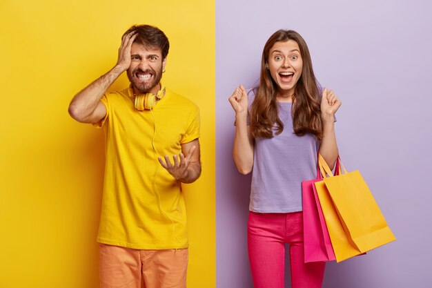 Cheerful female carries colorful shopping bags, rejoices new purchase, clenches fists with joy, annoyed husband feels angry with wife shopaholic, gestures with irritation