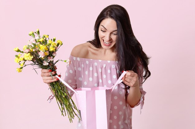 Cheerful female celebrates birthday, looks with happiness and surprise at gift bag, rejoices recieving present, holds beautiful flowers