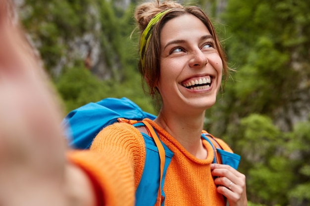 쾌활한 여성 탐험가는 녹색 숲에서 멋진 여행을하고 셀카를 만들기 위해 손을 뻗고 야외 배낭을 들고 서서 넓게 웃으며 옆으로 집중합니다. 사람들