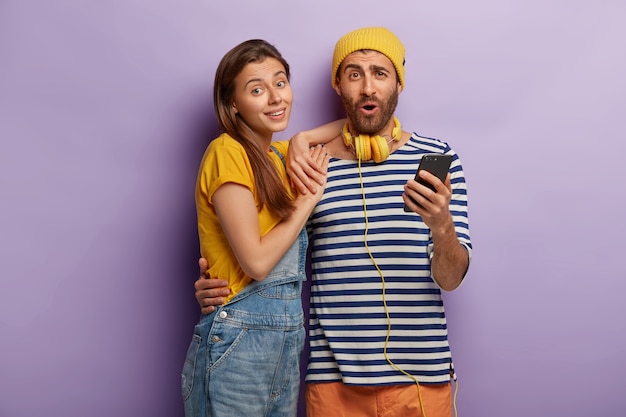 Free photo cheerful female and male embrace and use modern smartphone, dressed in stylish clothing, isolated on purple wall