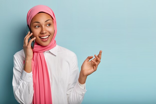 Cheerful female model discusses awesome news with close friend, holds smartphone near ear, looks right joyfully