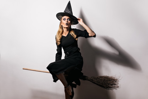 カーニバルの準備をしている長い黒のドレスと魔法の帽子の陽気な女性モデル。古いほうきで金髪の魔女の屋内ショット。