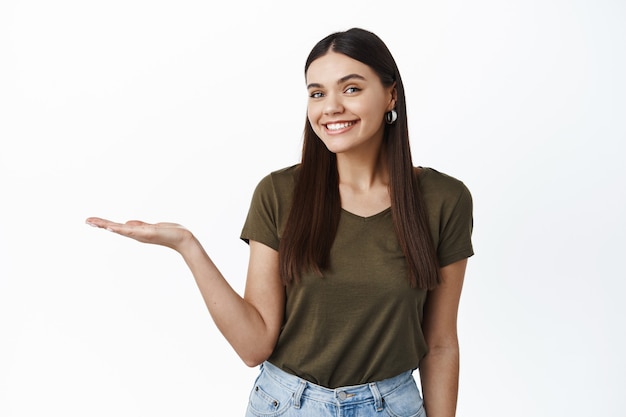 Free photo cheerful female model smiling at front excited while showing product on her hand, holding palm against white wall empty space