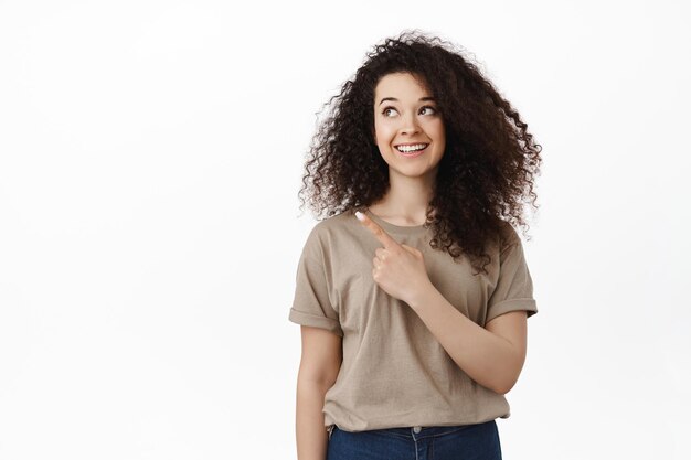 Cheerful female model with curly hairstyle pointing finger, looking at upper left corner with excited happy face, smiling white perfect teeth, standing in studio