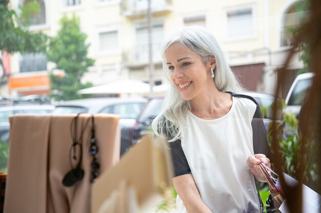 Cheerful female shopper looking at accessories in shop window, holding shopping bags, standing at store outside. Front view through glass. Window shopping concept