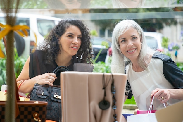 Cheerful female shoppers staring at accessories in shop window, holding shopping bags, standing at store outside. Front view through glass. Window shopping concept