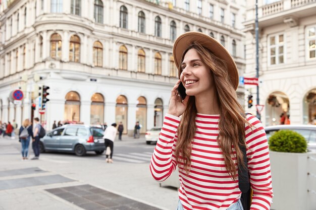 Cheerful female wanderer strolls in city, enjoys communication, keeps modern smartphone near ear, focused aside