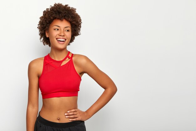 Cheerful fitness woman keeps hand on waist, being in good physical shape, wears red top, turns gaze away with happy smile, enjoys spending free time in gym