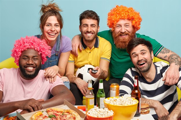 Веселые пятеро товарищей широко улыбаются, выражают положительные эмоции, радость, наблюдают за спортивным матчем, держат футбольный атрибут, радостно смеются, когда любимая команда побеждает соперника, съесть попкорн и выпить пива