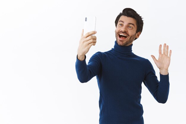 Cheerful friendly handsome bearded guy in high neck sweater holding smartphone in raised hand and smiling waving hand saying hi or hello waving palm having online conversation via videocall