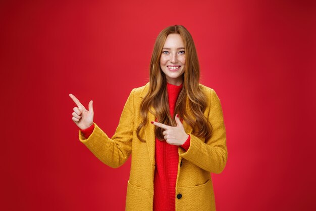 Веселая, дружелюбная и энергичная, услужливая рыжая девушка в желтом пальто показывает путь, указывая в левый верхний угол и широко улыбается с довольной ухмылкой, позируя в восторге на красном фоне.