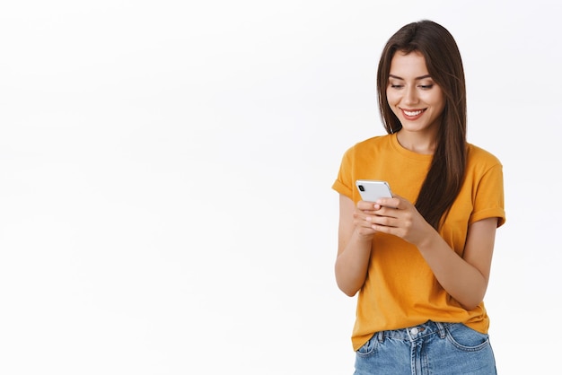 Веселая дружелюбно выглядящая женственная женщина в желтой футболке обменивается сообщениями, улыбаясь, счастливая и восхищенная, просматривая интернет с помощью смартфона, делая покупки в черную пятницу онлайн, размещая заказ на белом фоне