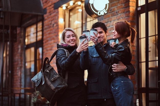 Cheerful friends making a toast with coffee while standing together near a cafe outside.