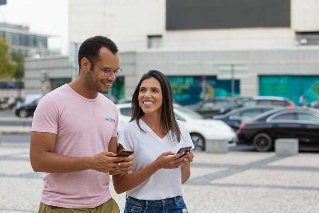 Cheerful friends strolling on street with smartphones