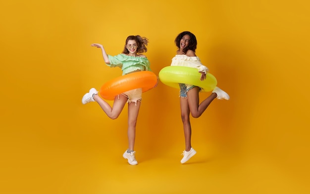 Cheerful friends woman dressed in summer clothes jumping with rubber ring on yellow background