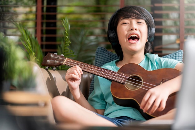 Cheerful fun asian child boy singing loud play ukulele with exiting and energy asian male son make loud funny noise practicing ukulele online lesson at home stay home concept