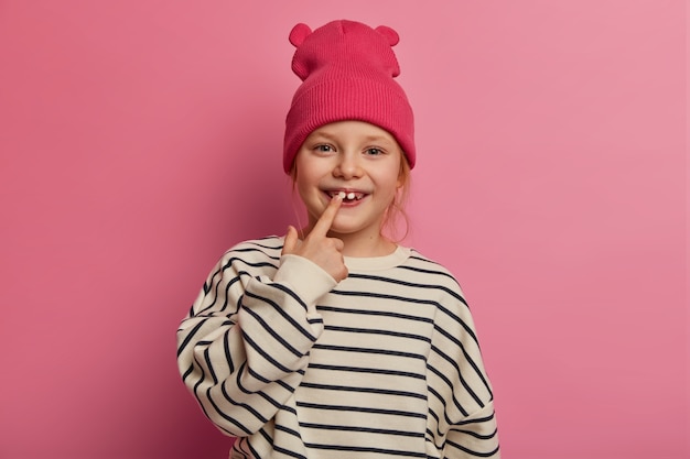 Cheerful funny little girl points at her teeth, cares about oral hygiene, dressed in fashionable clothes, has healthy skin, boasts of adult tooth to friends on playground, isolated on pink pastel wall