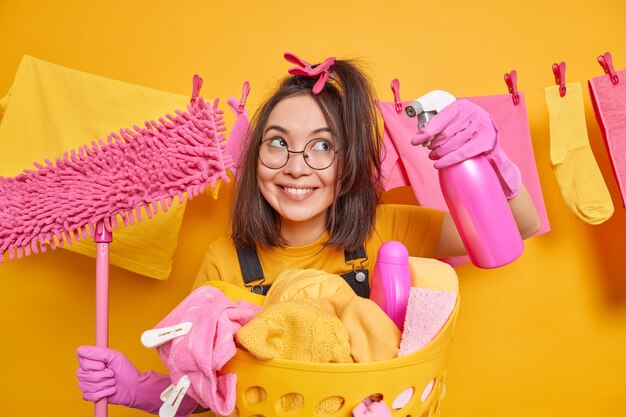 Cheerful funny millennial girl wears round spectacles and rubber gloves poses with cleaning supplies does laundry at home poses against clothesline hanging over yellow wall