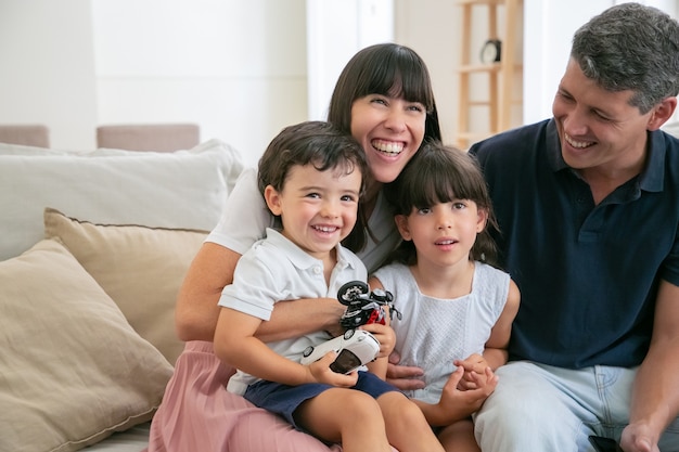 Cheerful funny parents and two kids watching funny movie at home, sitting on couch in living room and looking away and laughing.