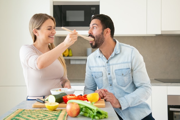 陽気な面白い若いカップルが一緒に夕食を作って、キッチンで新鮮な野菜をカットします。彼女のボーイフレンドに大きなスプーンで食べ物のスライスを与えて味わう女性。家族の料理のコンセプト