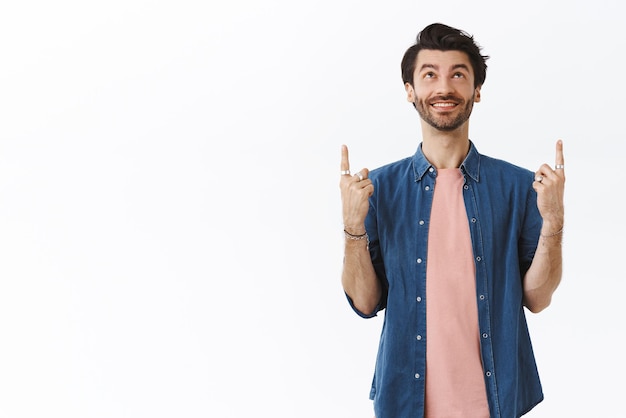 Free photo cheerful gay man with beard hipster hairstyle wearing jewelry modern outfit looking and pointing up amused smiling happy checking out advertisment saw awesome promo standing white background