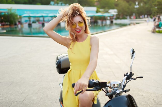 Cheerful ginger woman in yellow glasses and vintage dress sitting on stylish motorbike. Vacation mood. Sunny summer day. Red lips.