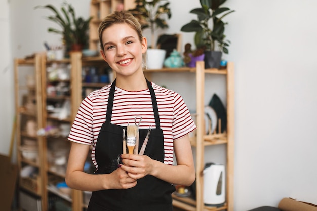 Free photo cheerful girl in black apron and striped t-shirt holding pottery tools in hands happily looking in camera at pottery studio