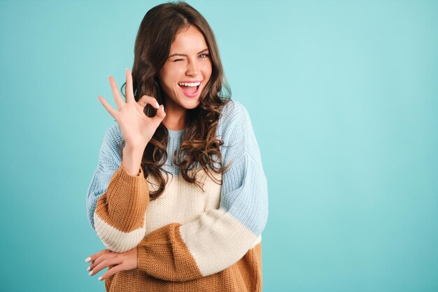 居心地の良いセーターの陽気な女の子は、青い背景の上にカメラで大丈夫なジェスチャーとウインクを喜んで示しています