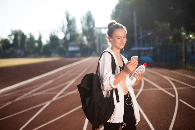 Cheerful girl in earphones happily using cellphone with bottle of water in hand and backpack on shoulder while spending time on racetrack of stadium